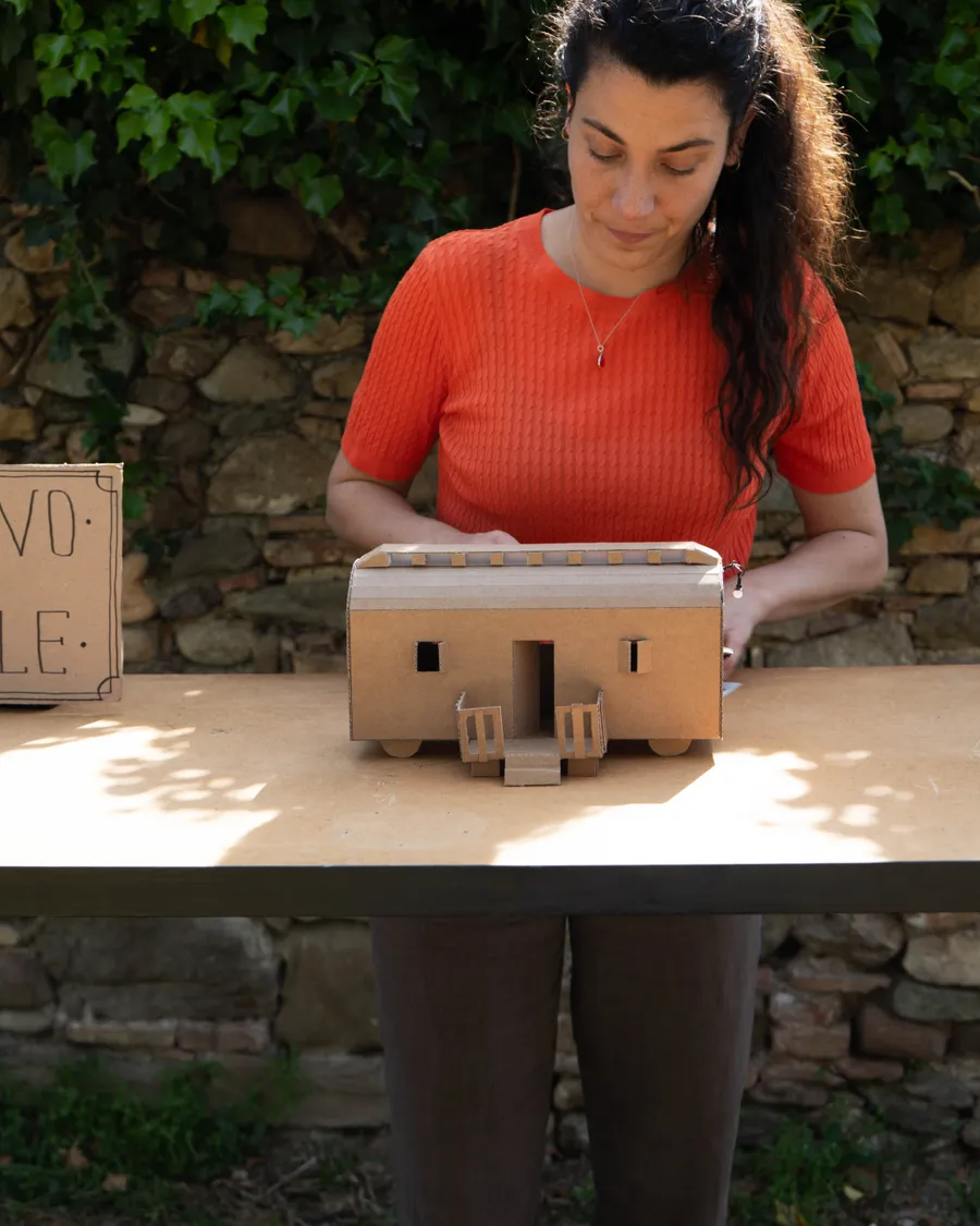 Mujer con jersey naranja observando una maqueta de cartón de una casa sobre una mesa al aire libre, junto a un cartel de la instalación, con un muro de piedra al fondo.