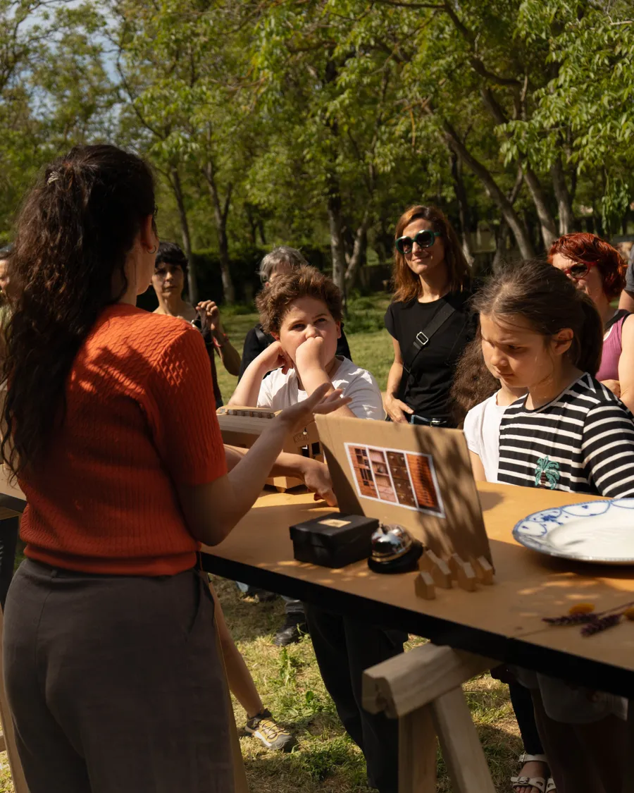La misma mujer explicando la instalación Archivo de lo posible a un grupo de niños y adultos alrededor de una mesa con maquetas y objetos.