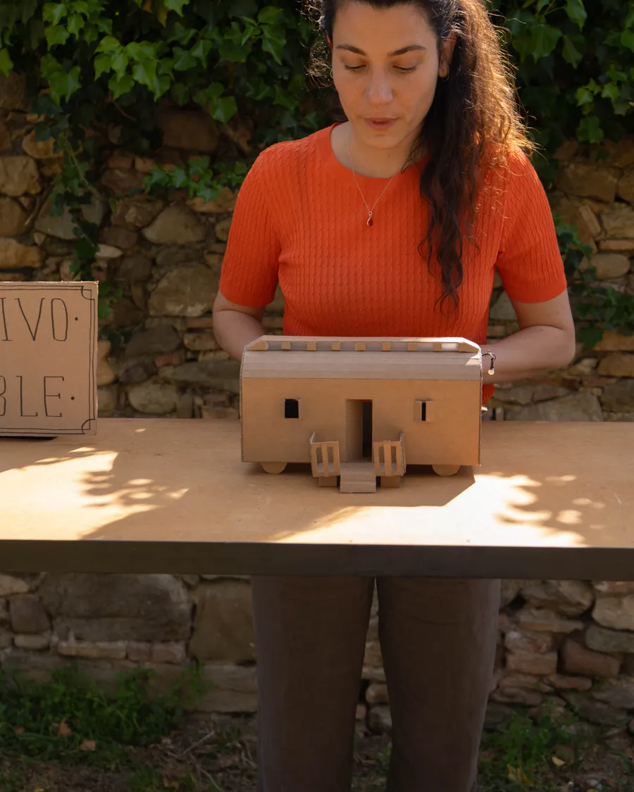 Primer plano de la misma mujer mirando la maqueta de cartón de la casa, con el cartel de la instalación desenfocado al fondo.