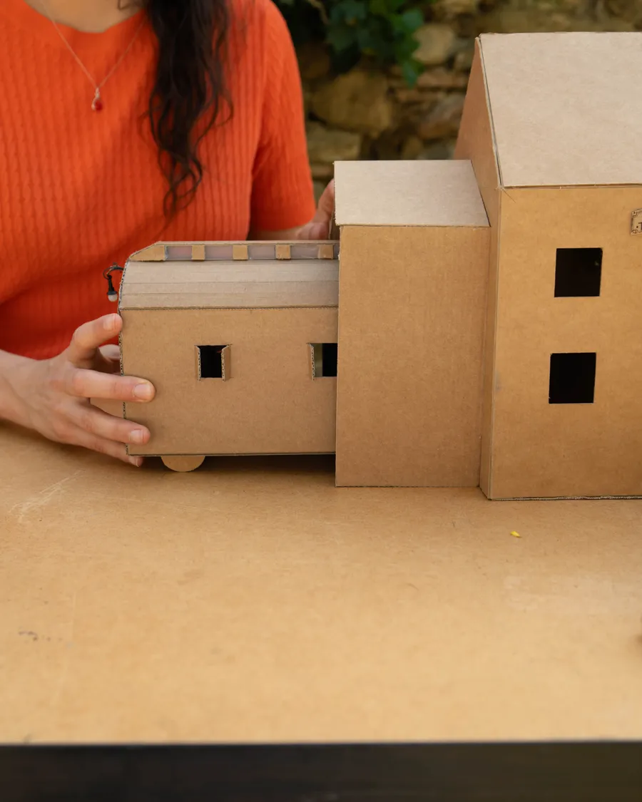 Detalle de varias maquetas de cartón: una casa pequeña con ruedas y dos edificios de dos plantas con ventanas recortadas.