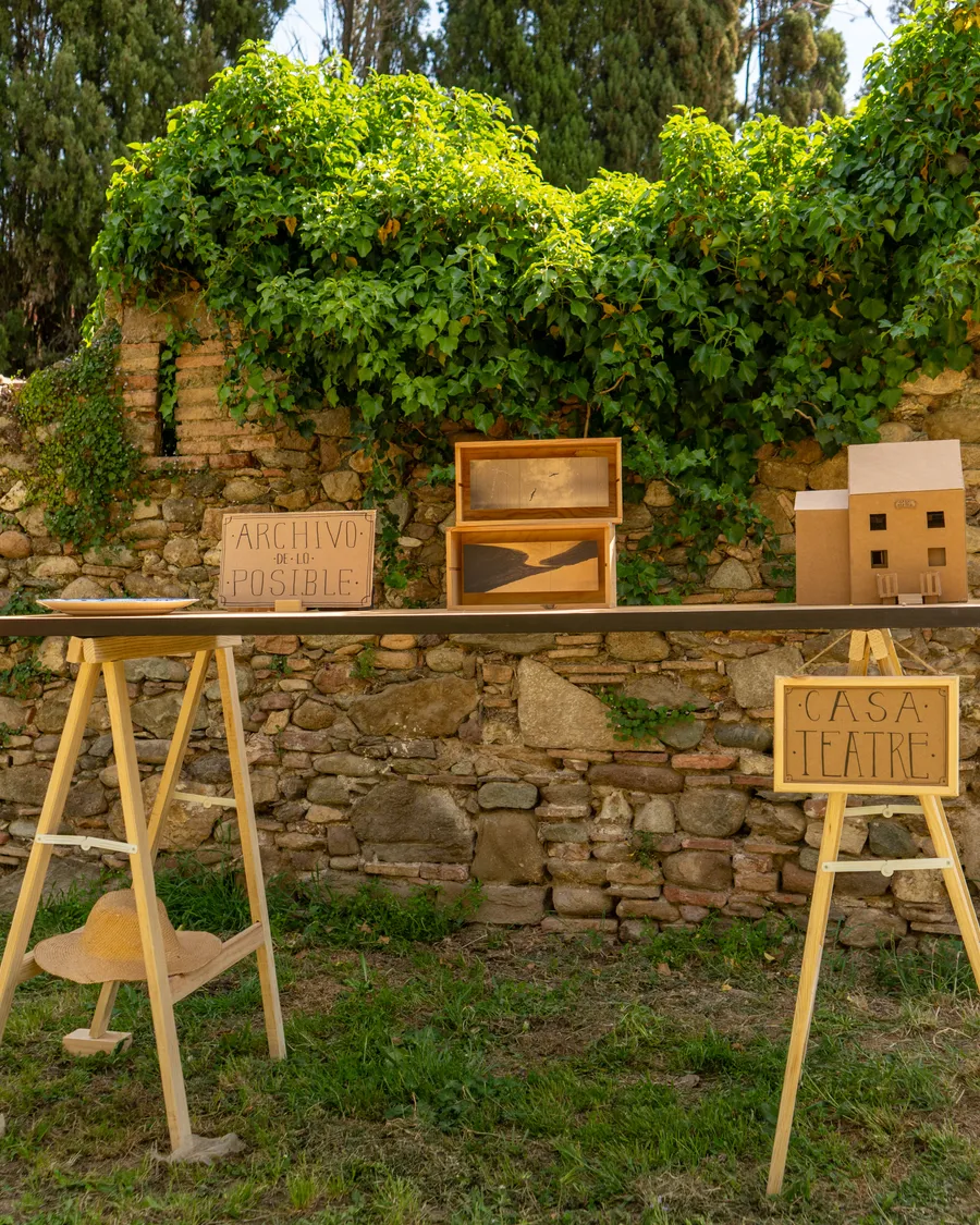 Instalación al aire libre con el cartel Archivo de lo posible, una caja de luz con una fotografía y las maquetas de cartón de Casa Teatre junto a un muro de piedra.