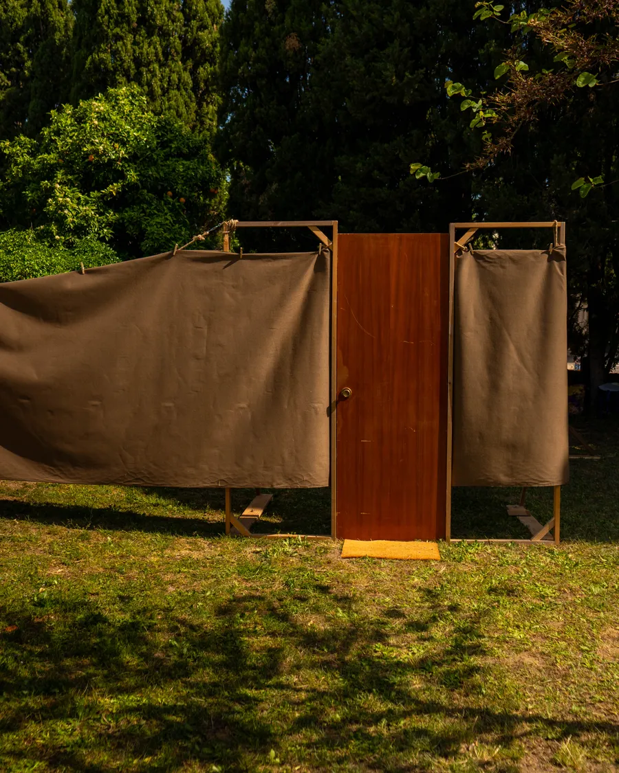 Estructura escénica al aire libre con cortinas de tela marrón y una puerta de madera entre árboles, parte de la instalación Casa Teatre.