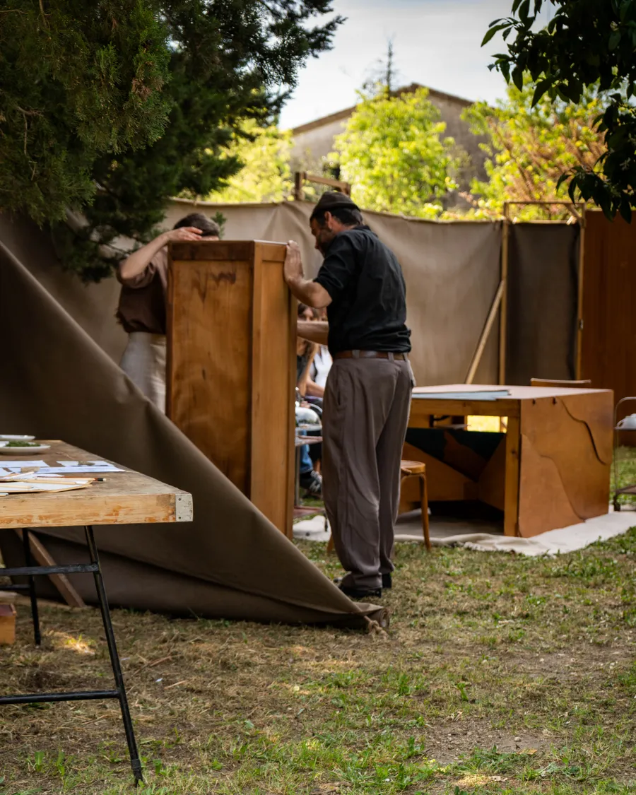 Dos personas montando una estructura de madera y tela para la instalación al aire libre, con mesas y materiales alrededor.