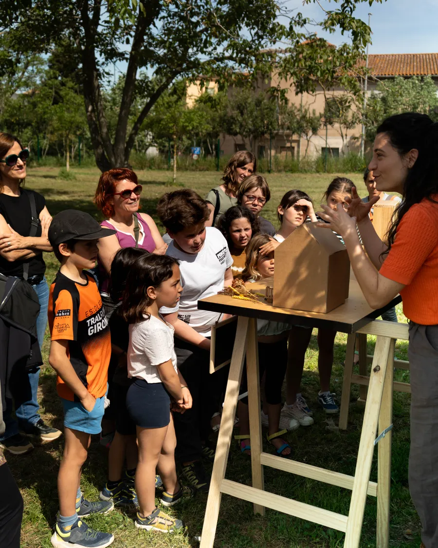 Grupo de niños y adultos observando una maqueta de cartón mientras una mujer con jersey naranja les explica el proyecto al aire libre.