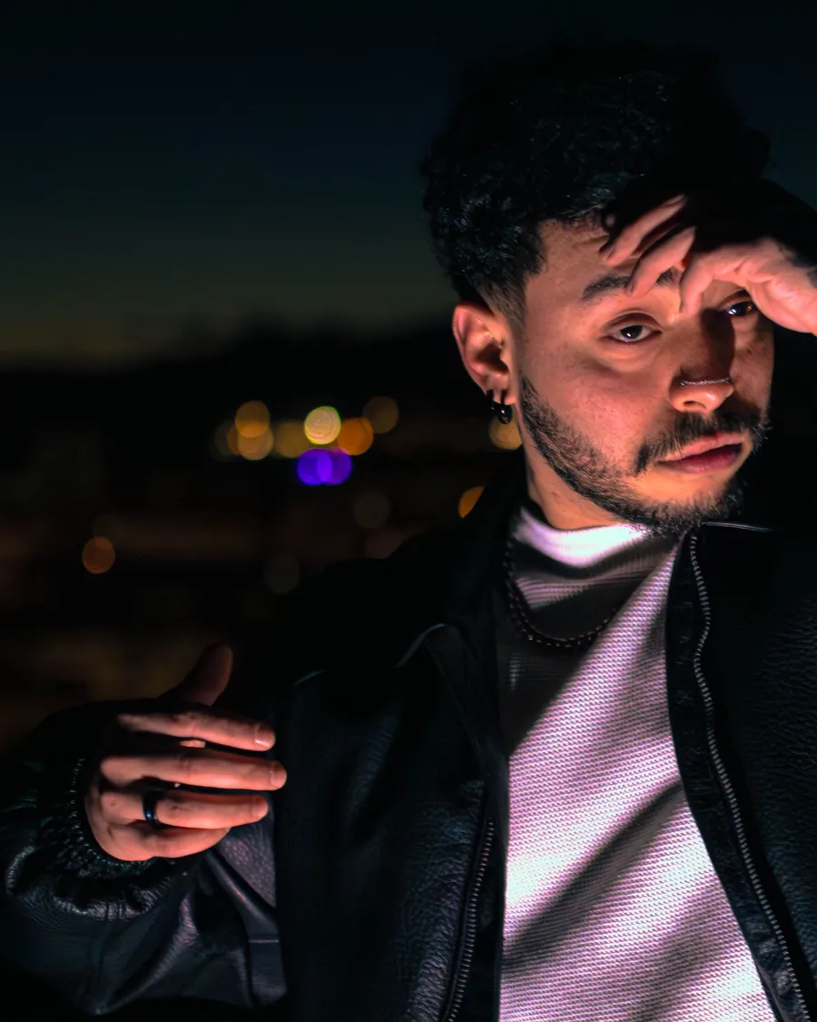 Retrato nocturno de JoshI4X con la mano cerca del rostro, iluminado con luces de neón azules y violetas y la ciudad de fondo.