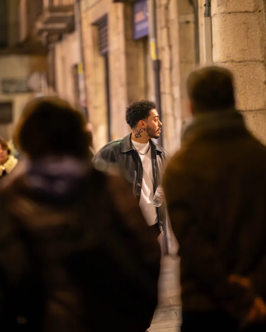 JoshI4X caminando de noche por una calle de arquitectura de piedra, rodeado de otras personas desenfocadas en primer plano.